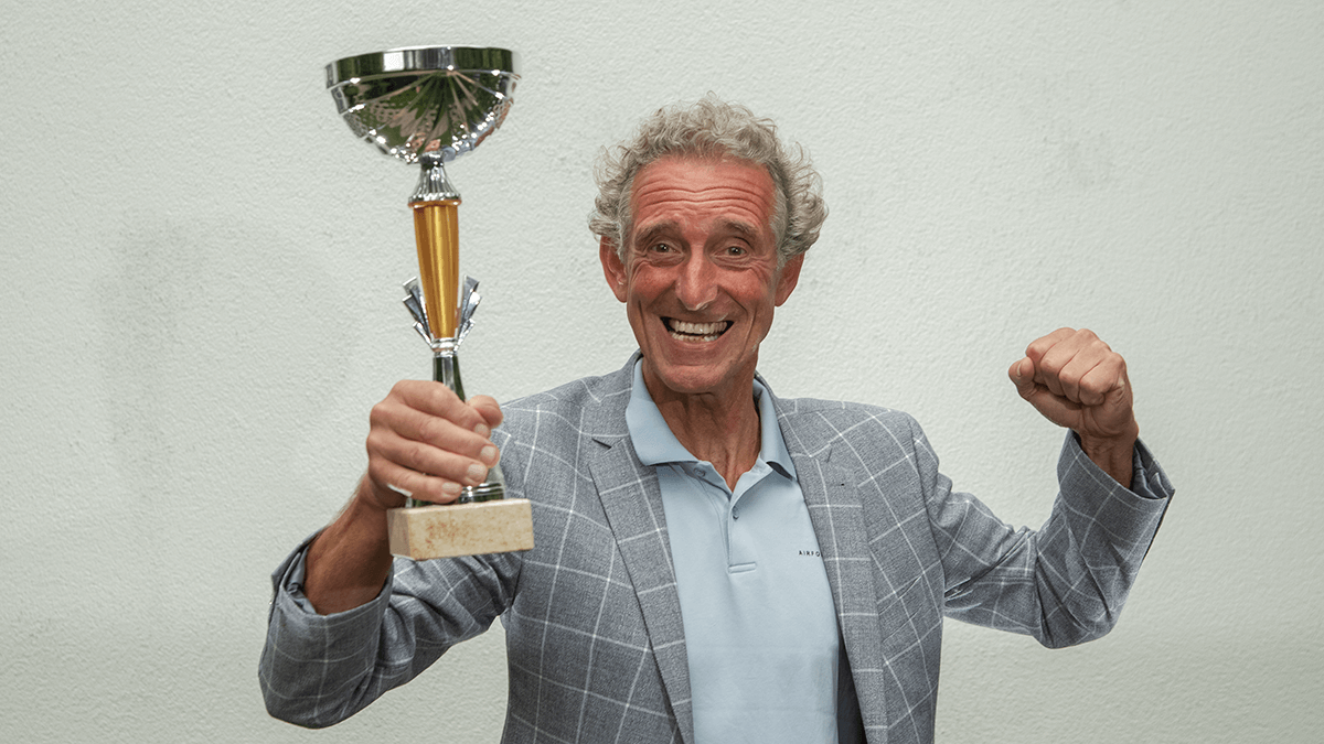 Foto van coach Cor van Ruijven van CvR VISIE. Op de foto draagt Cor een licht grijs geruiten colbert en een polo. Cor van Ruijven staat blij op de foto en heeft een bokaal in zijn hand.